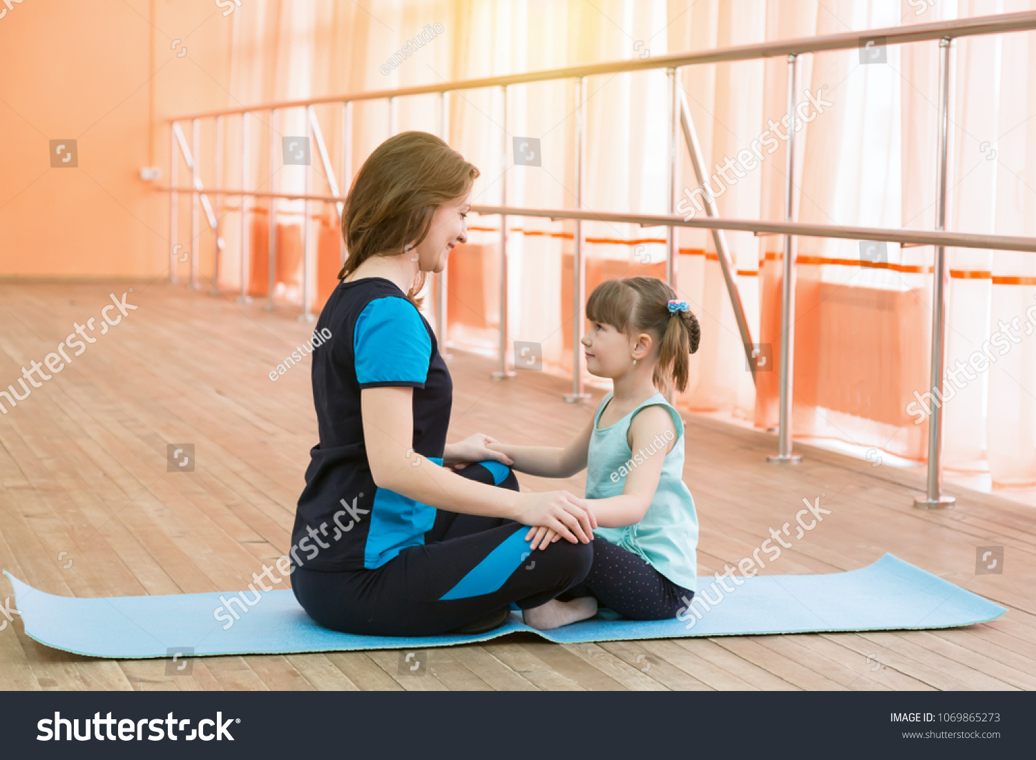 Doing sports with moms and daughters. Young woman and little girl are sitting opposite each other on a sports carpet. #1069865273