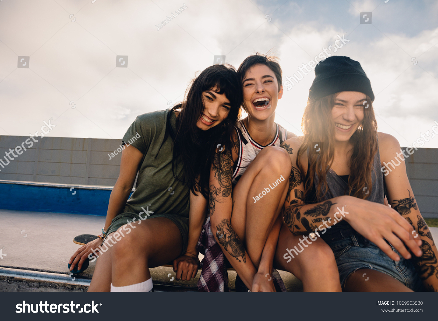 Shot of three smiling girls hanging out at skate park. Group of women friends sitting outdoors at skate park and laughing. #1069953530