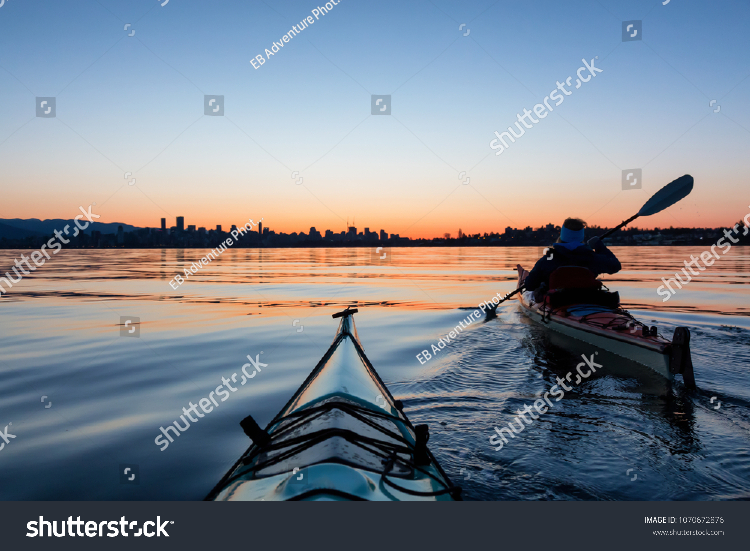 Adventurous Girl Sea Kayaking during a vibrant winter sunrise with City Skyline in Background. Taken in Downtown Vancouver, British Columbia, Canada. Concept: Adventure, Holiday, Vacation, Sport #1070672876