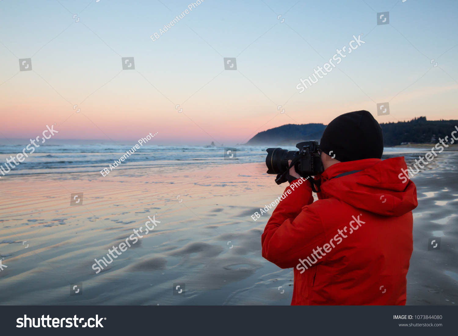 Photographer with a camera is standing on the sandy beach during a vibrant and colorful winter sunrise. Taken in Canon Beach, Oregon Coast, United States of America. #1073844080