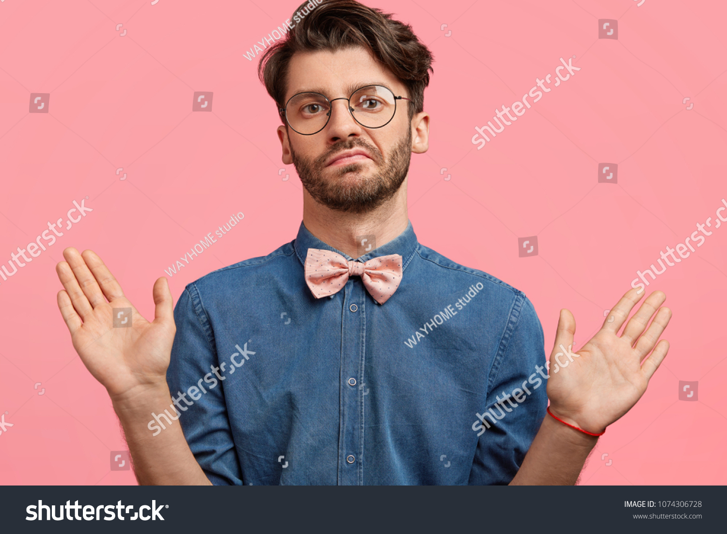 I am not guilty! Fashionable bearded guy being hesitant or shows his non participation in something, has bristle, wears stylish denim shirt, isolated over pink studio background. Body language #1074306728