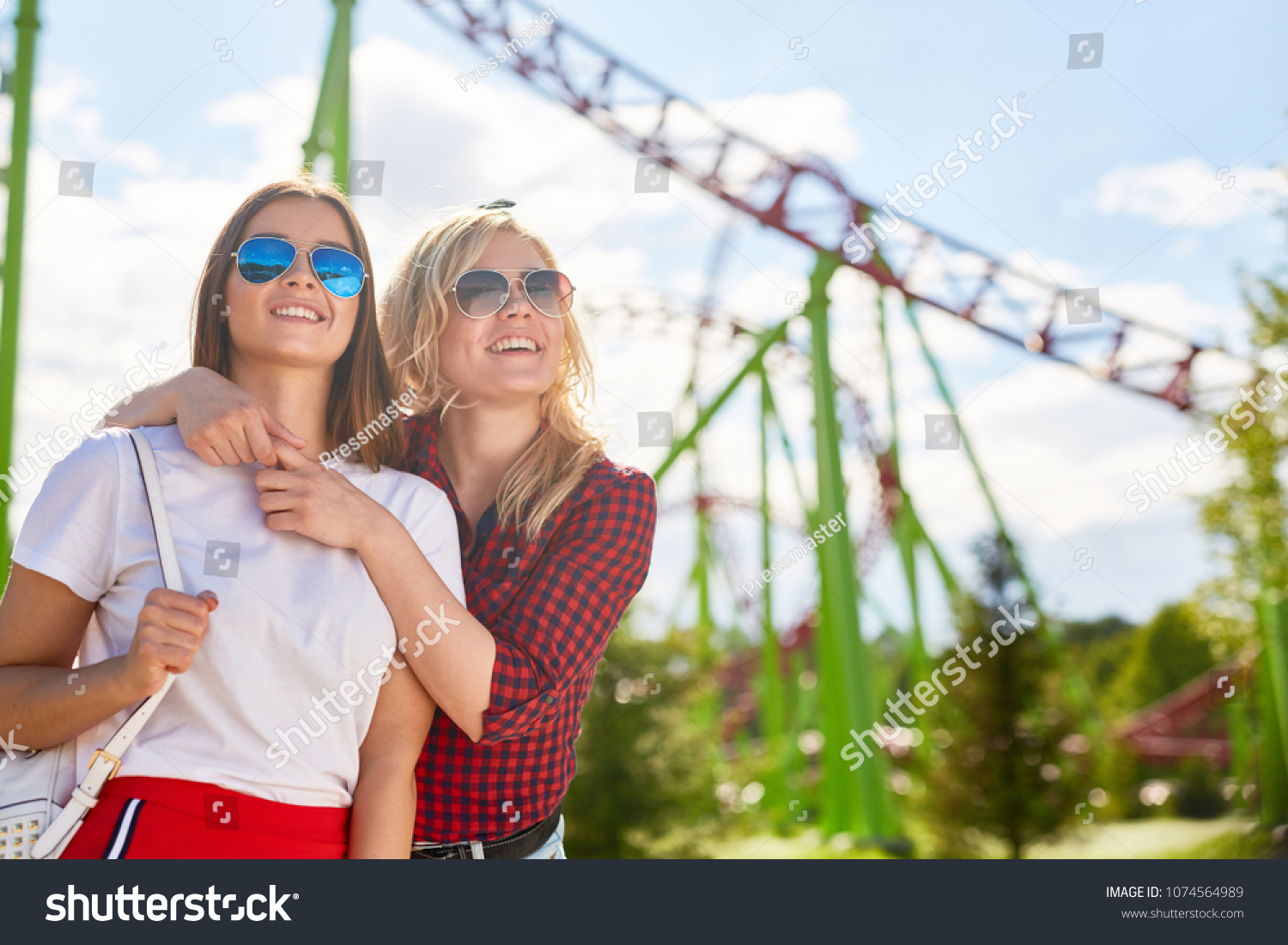 Two affectionate girls in sunglasses looking at one of amusements while spending time in theme park on summer weekend #1074564989
