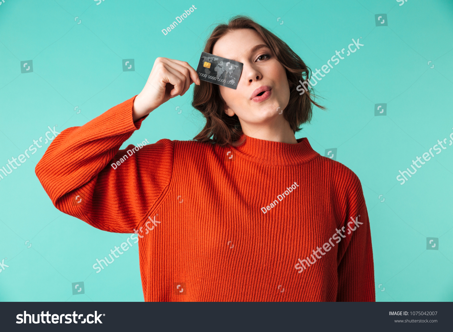 Portrait of a pretty young woman dressed in sweater holding credit card at her face isolated over blue background #1075042007