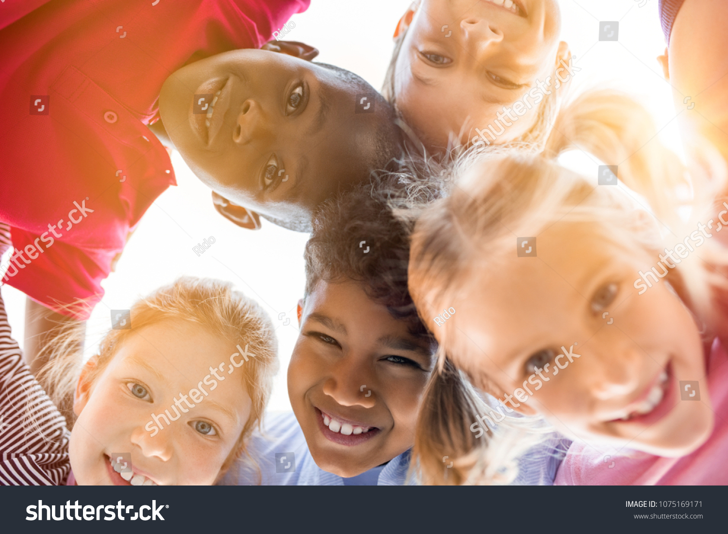 Portrait of happy kids in circle looking down and embracing. Group of five multiethnic friends outdoor looking at camera and smiling. Closeup face of children looking at camera together at park. #1075169171