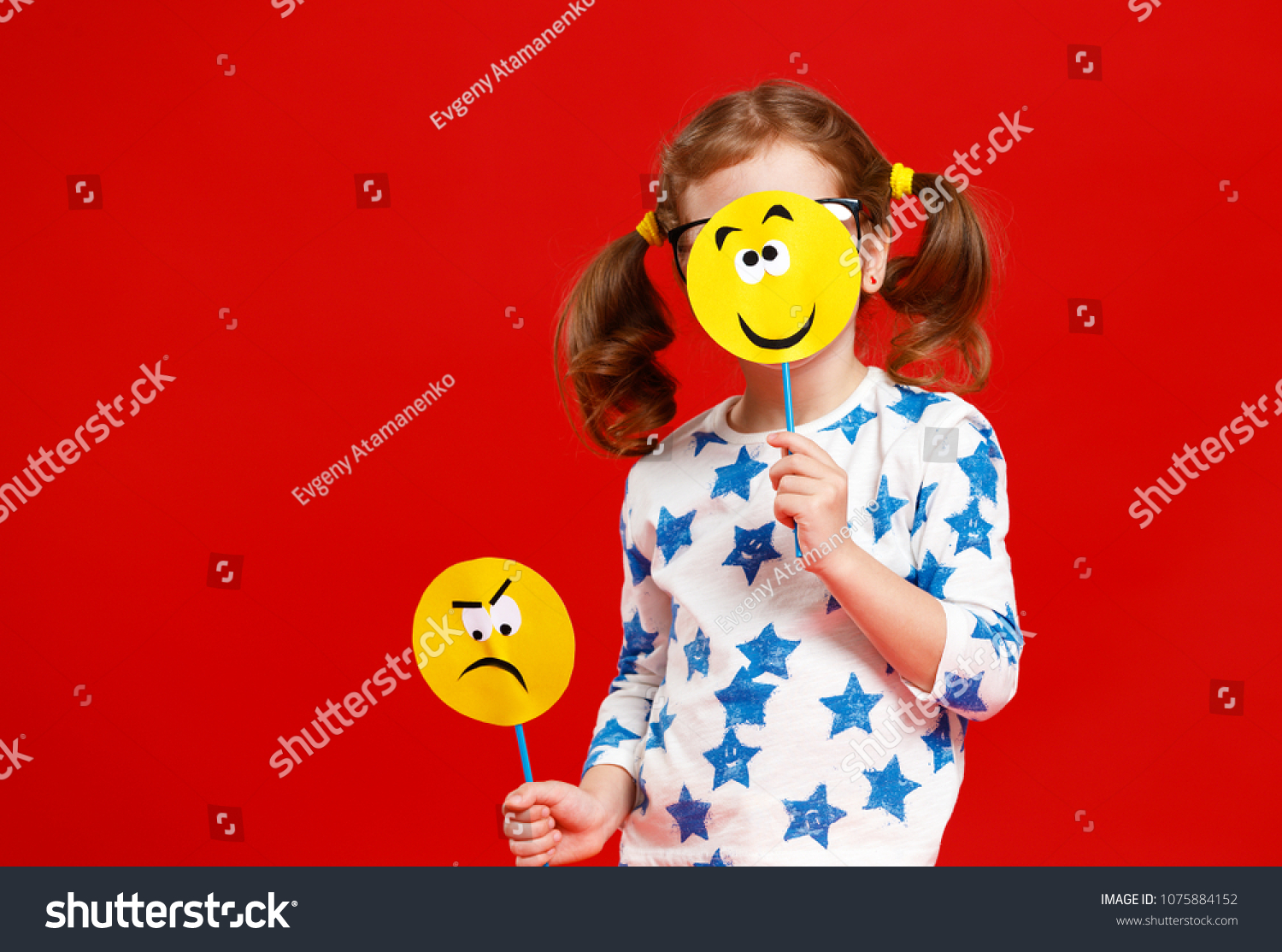 concept of children's emotions. child girl chooses between a sad and joyful smile on  colored red background
 #1075884152