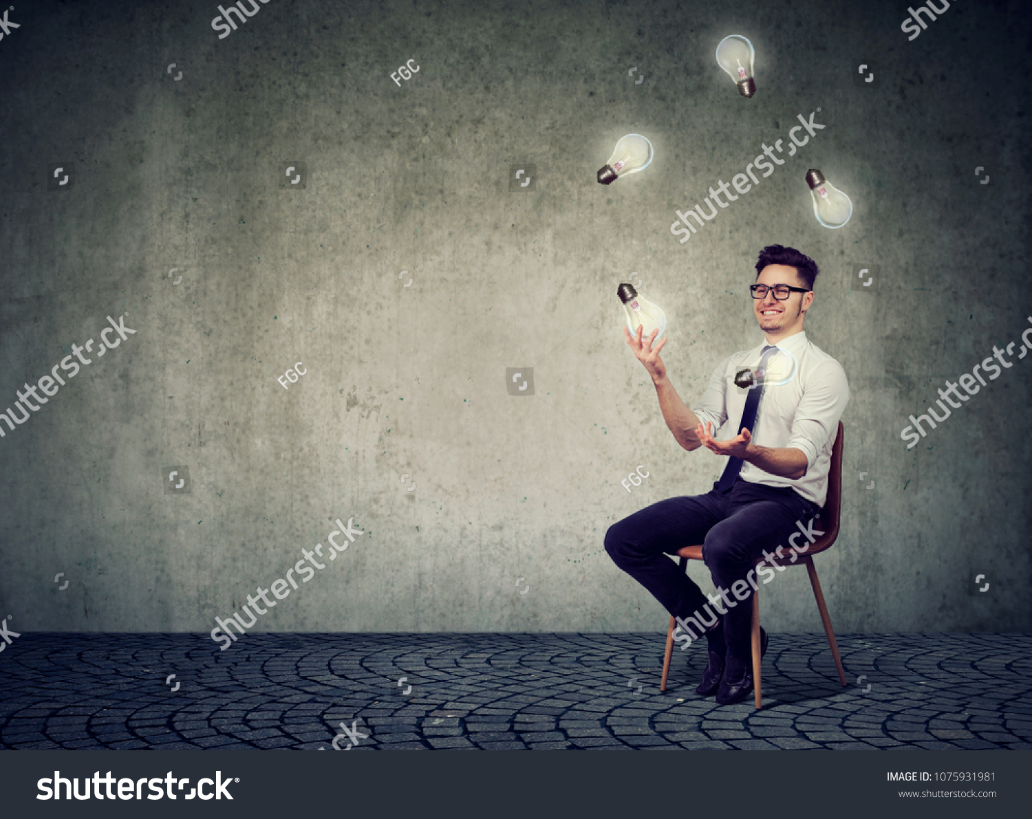 Happy smart man sitting on chair against gray concrete wall and juggling with light bulbs having many brilliant ideas. #1075931981