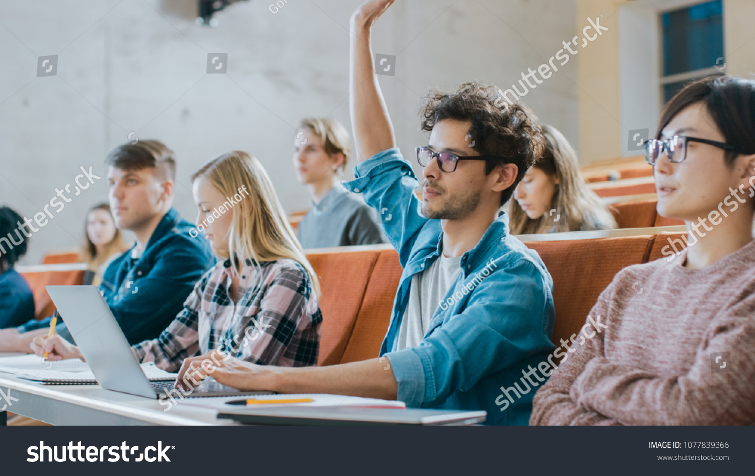 Handsome Hispanic Student Uses Laptop while Listening to a Lecture at the University, He Raises Hand and Asks Lecturer a Question. Multi Ethnic Group of Modern Bright Students. #1077839366