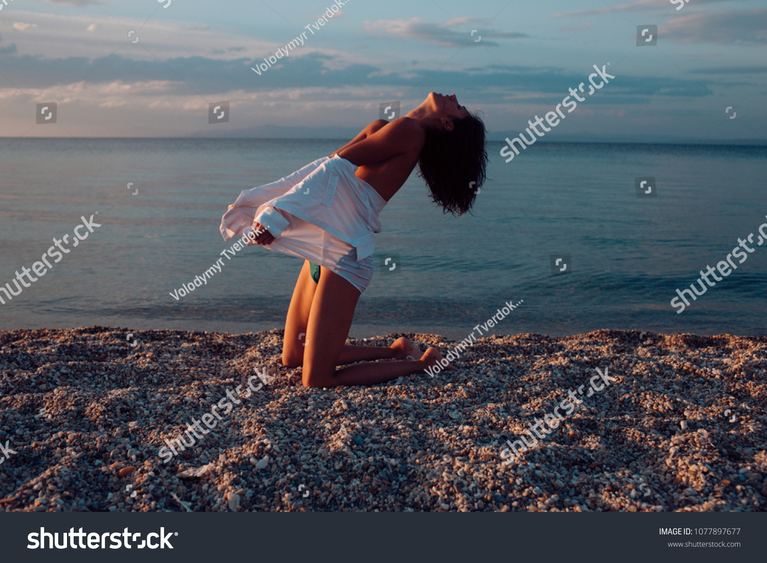 Attractive young lady suntanning nude, nudist, enjoy last sun rays. Woman stands on knees on beach in evening. Girl sexy, topless, naked breasts with wild hair at seashore at sunset. Erotic concept. #1077897677