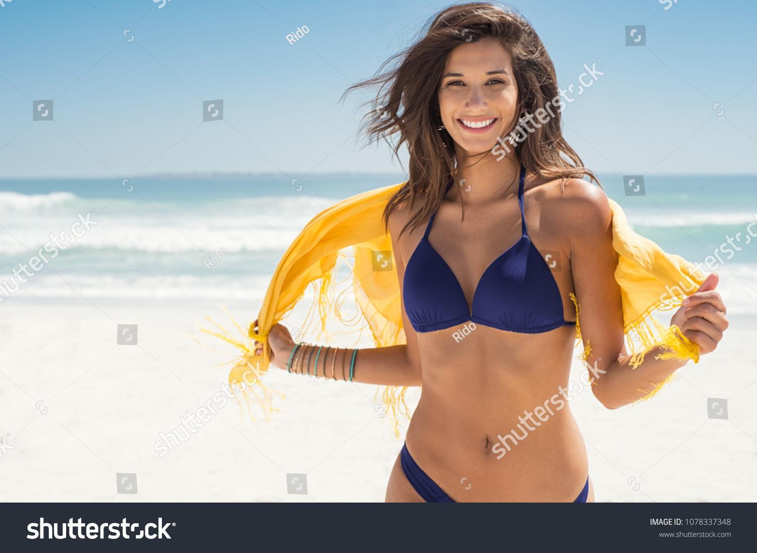 Happy woman smiling and having fun at beach. Summer portrait of young beautiful girl running on beach with a yellow scarf. Latin girl laughing and looking at camera with joy.  #1078337348