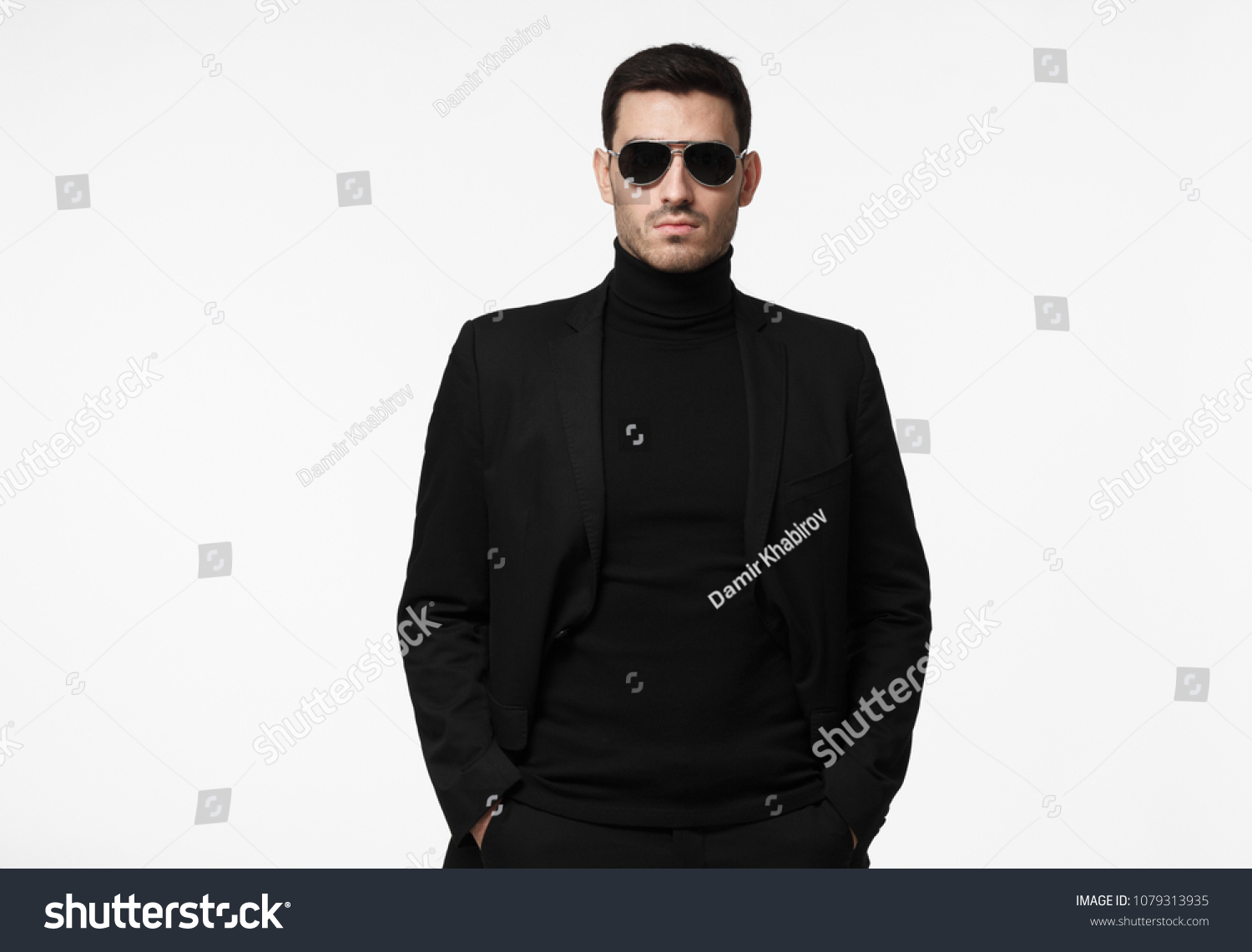 Half-length portrait of young handsome European guy isolated on white background dressed in total black with turtleneck, sunglasses and jacket, looking serious and suspicious, reserved and reticent #1079313935