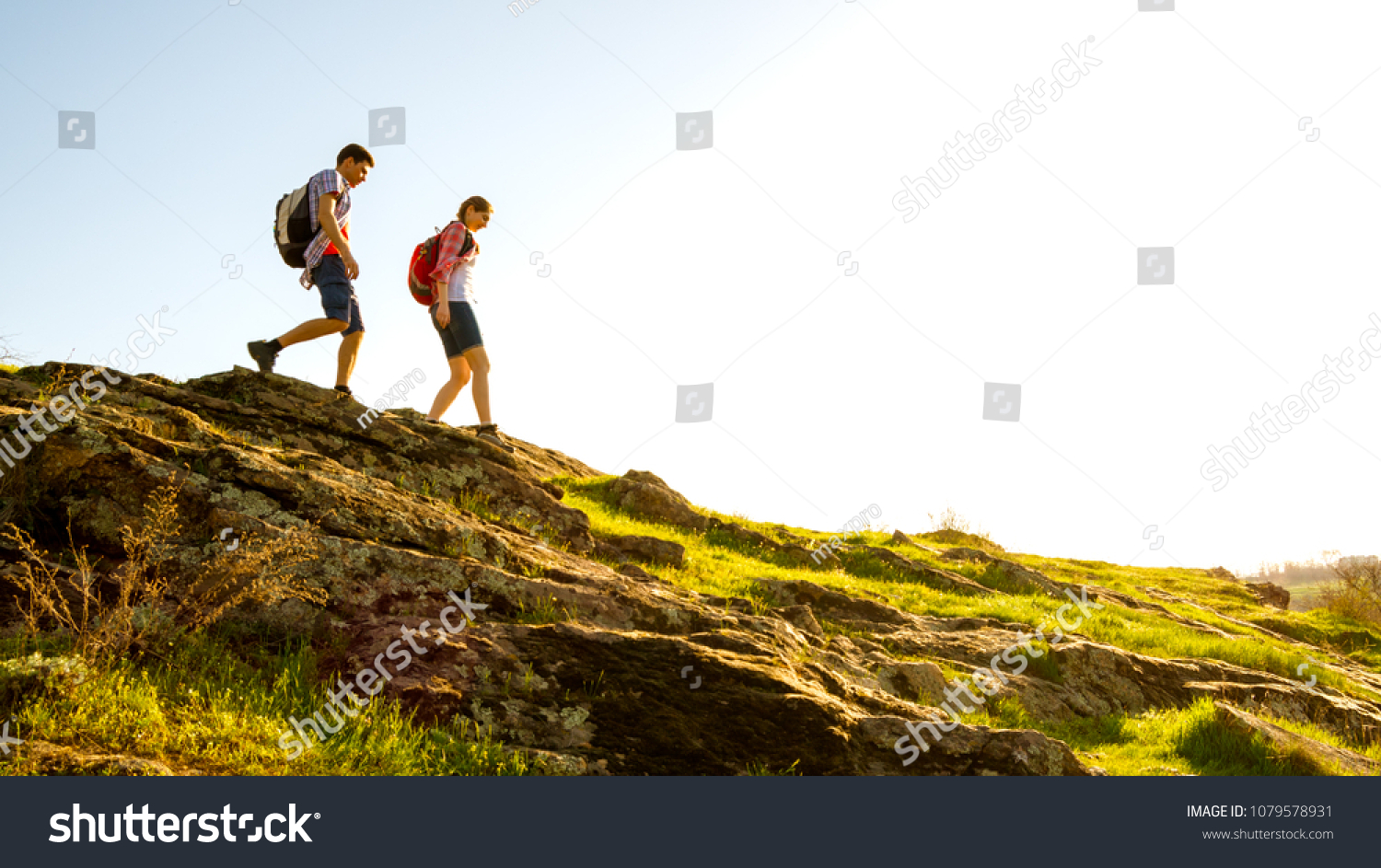 Couple of Young Happy Travelers Hiking with Backpacks on the Beautiful Rocky Trail at Warm Sunny Evening. Family Travel and Adventure Concept. #1079578931