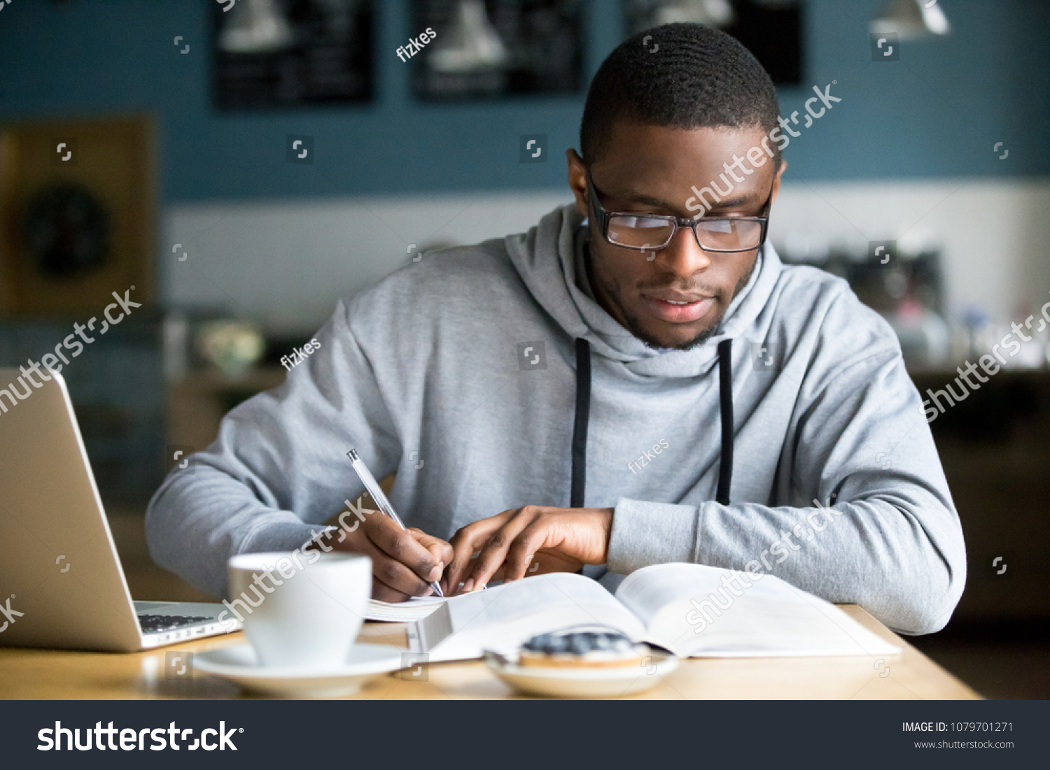 Focused millennial african american student in glasses making notes writing down information from book in cafe preparing for test or exam, young serious black man studying or working in coffee house #1079701271