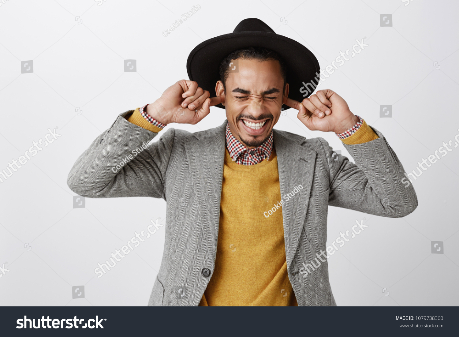Using natural earplugs not to hear spoilers. Good-looking happy dark-skinned guy in classy hat and stylish outfit smiling with closed eyes, holding index fingers in ears, waiting for surprise #1079738360