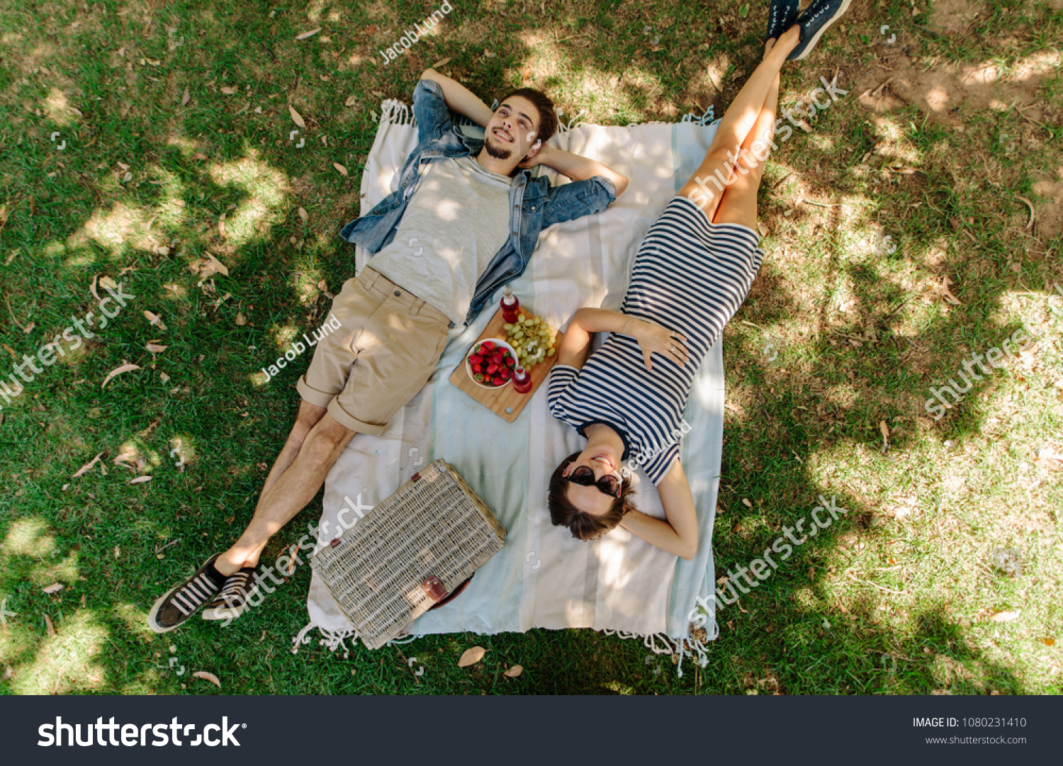 View from above of relaxed man and woman lying down at the park with hand basket, appetizing snacks and beers on blanket. Couple on picnic lying on blanket in grass outdoors. #1080231410