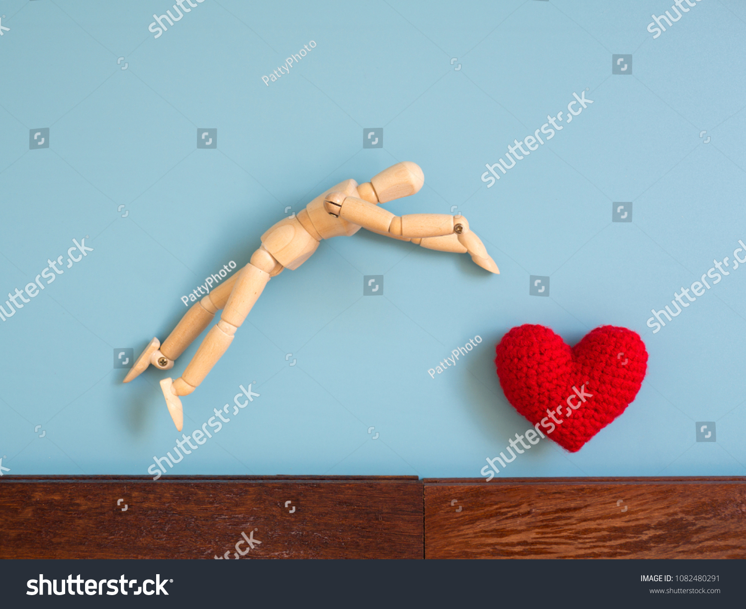 Wooden puppet jumping from the wooden floor in the air to catch the red heart floating in the sky. Wooden puppet try to jump to catch love. Concept of effort and love #1082480291