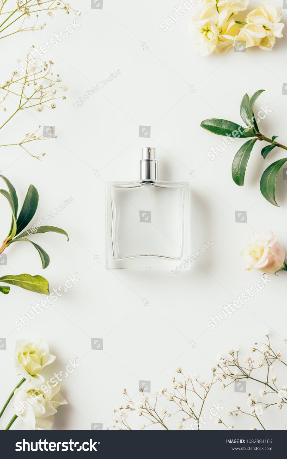 top view of bottle of perfume surrounded with flowers and green branches on white #1082884166