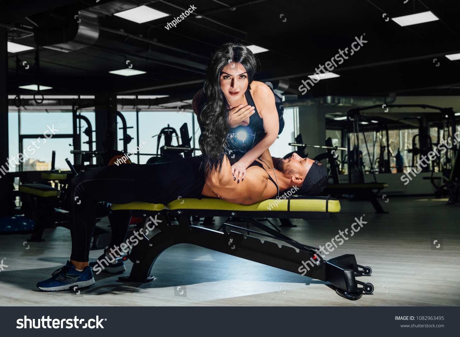 man doing barbell bench press and holds a girl instead of the bar in his arms. self-weight exercise at gym. copy space. beautiful fitness model amazing show training couple. guy has a bristle and cap. #1082963495