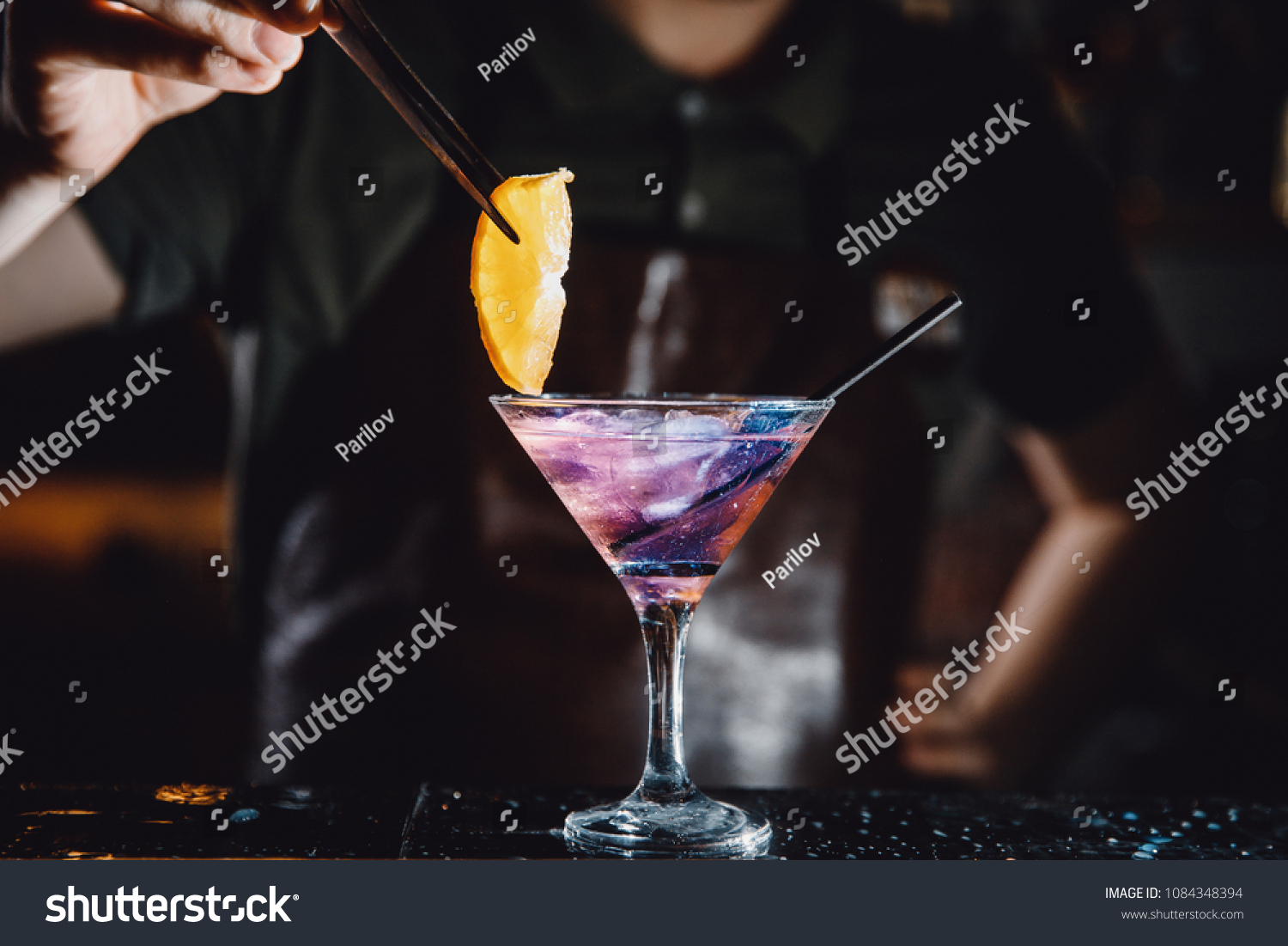 Barman prepares cocktail with orange and martini purple color on bar with alcohol. Uses tongs for decoration. Dark background. #1084348394