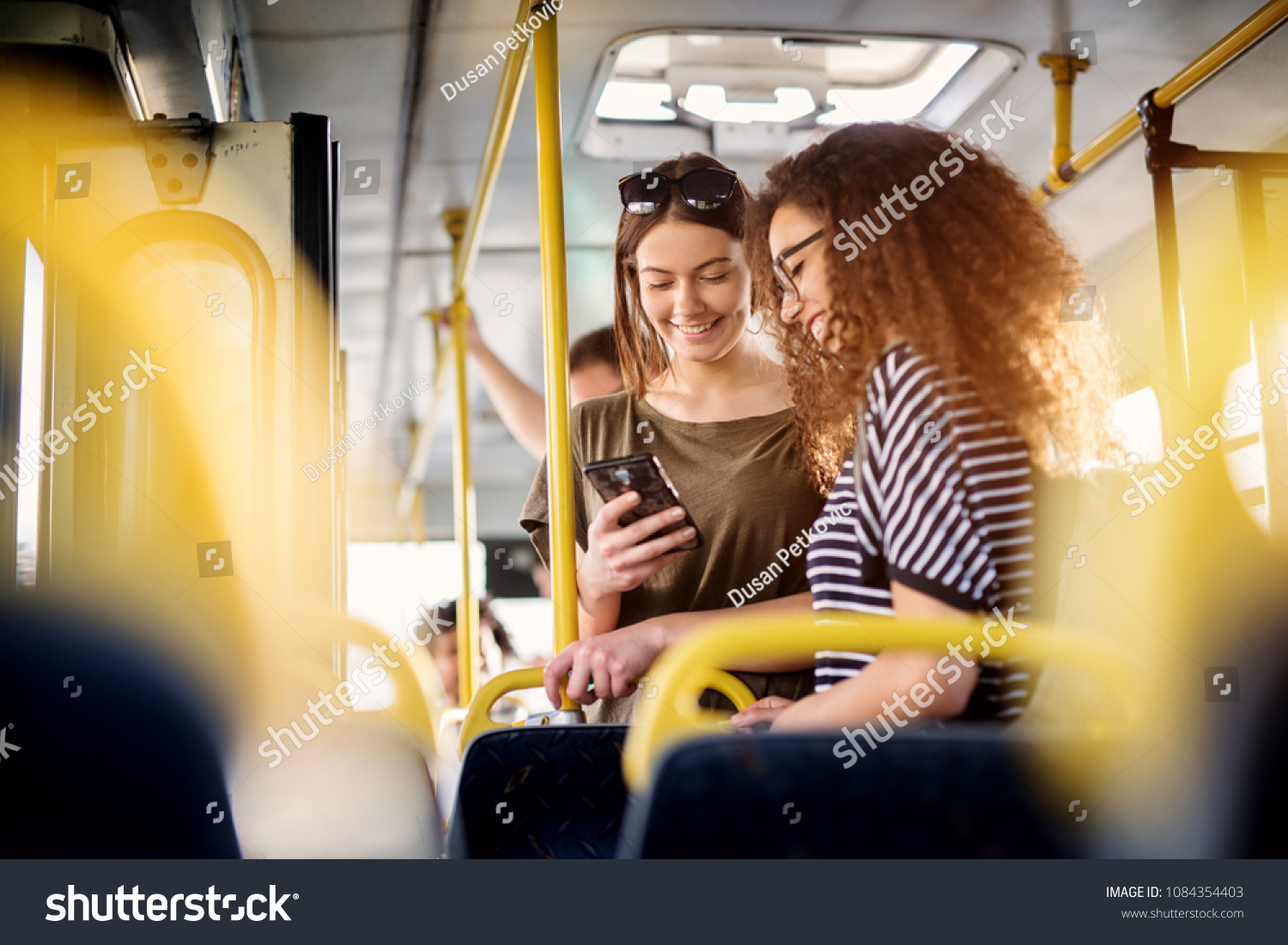 Two cheerful pretty young women are standing in a bus and looking at the phone and smiling while waiting for a bus to take them to their destination. #1084354403