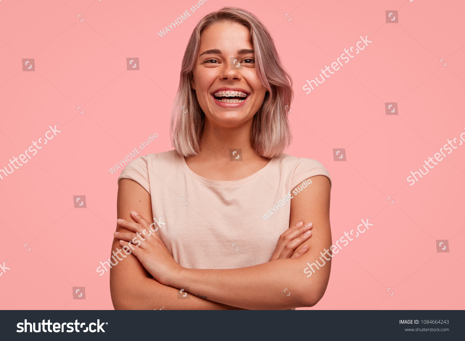 People, happiness and facial expressions concept. Pretty young woman with broad shining smile, keeps hands crossed, being in high spirit, wears braces on teeth, poses alone against pink background #1084664243