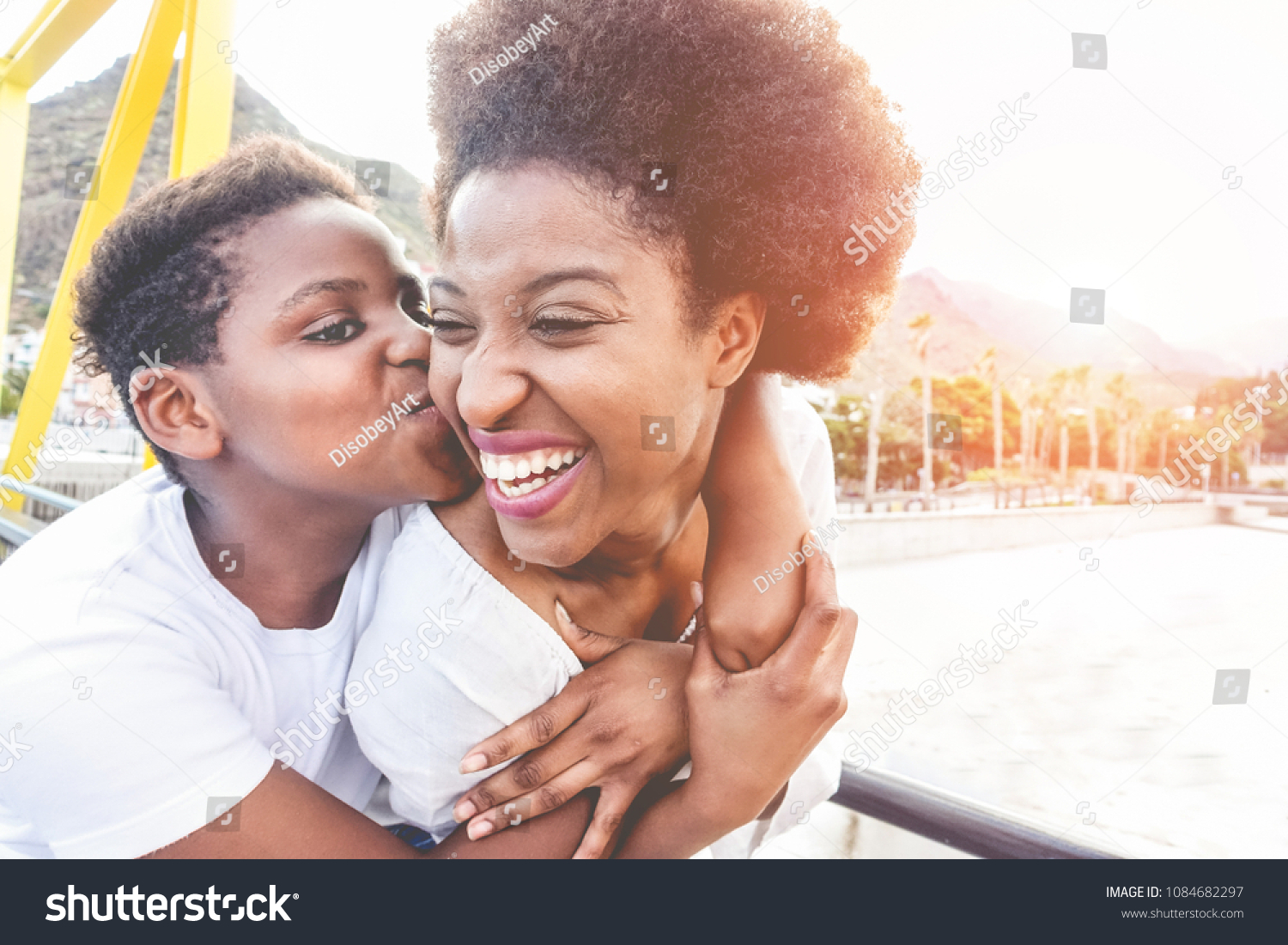 Happy young mother having fun with her child in summer sunny day - Son kissing his mum outdoor with back sun light - Family lifestyle, motherhood, love and tender moments concept - Focus on woman face #1084682297