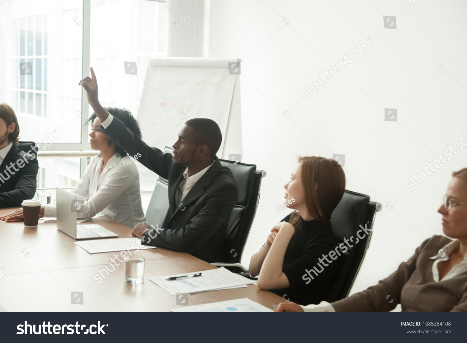 African businessman in suit raising hand at corporate diverse group meeting, black employee voting as volunteer asking question at business training sitting at conference table with multiracial team #1085354108