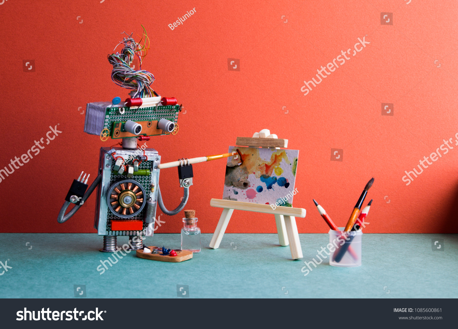 Talented robot artist paints an abstract picture with watercolors. Wooden easel and artist's tools palette, pencils case. Advertising poster studio school of visual arts interior.  #1085600861
