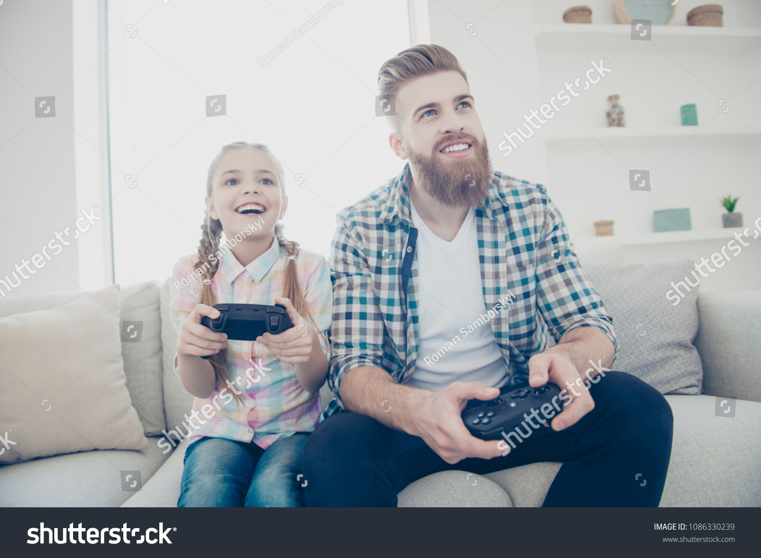 Cheerful joyful excited stylish trendy father and daughter holding joy-sticks in hands playing video game sitting on couch indoor in living room enjoying free time wearing casual outfit front view #1086330239