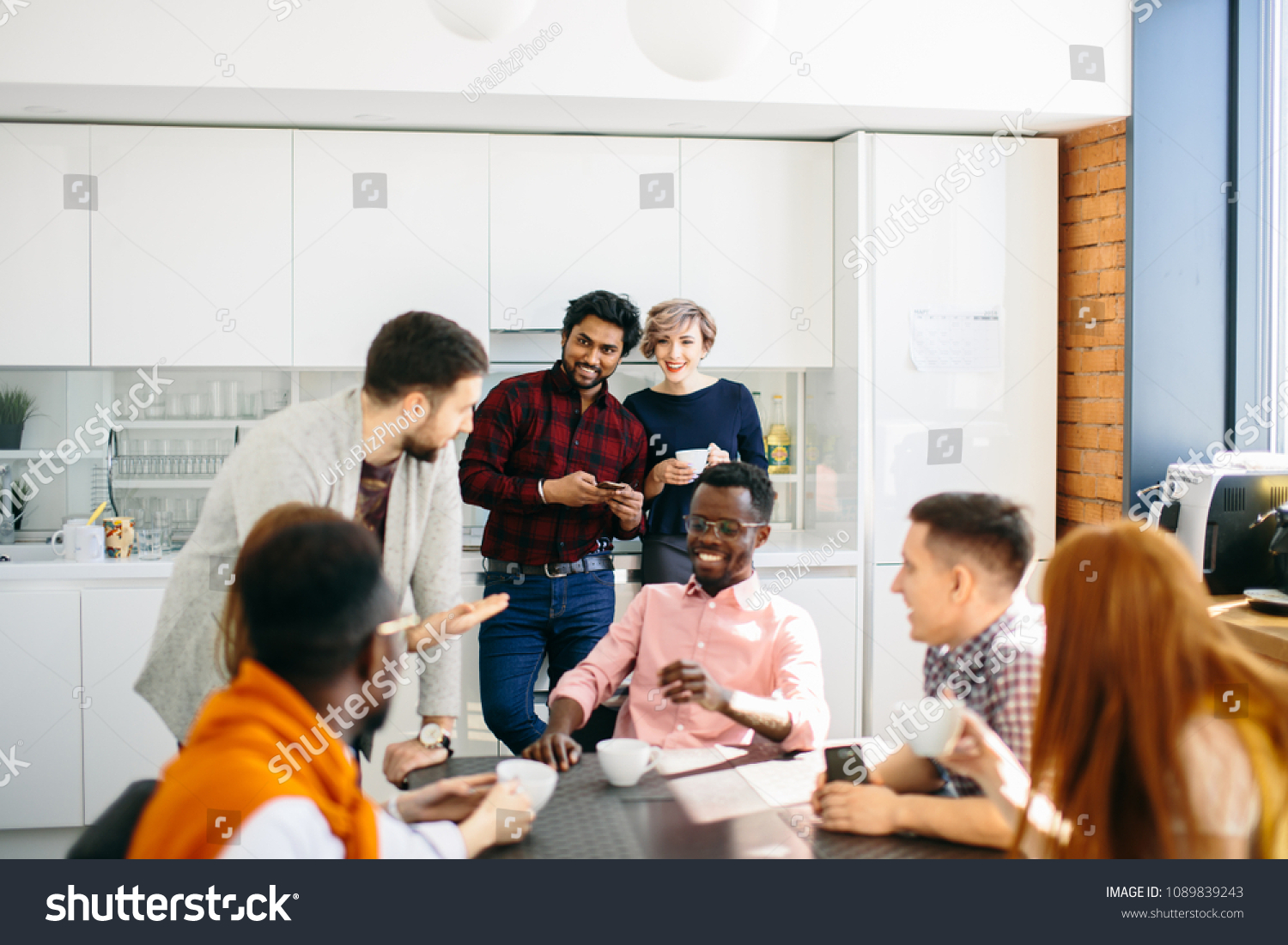 cheerful young Indian man and elegant Caucasian woman standing and looking at colleagues who are sitting at the table #1089839243