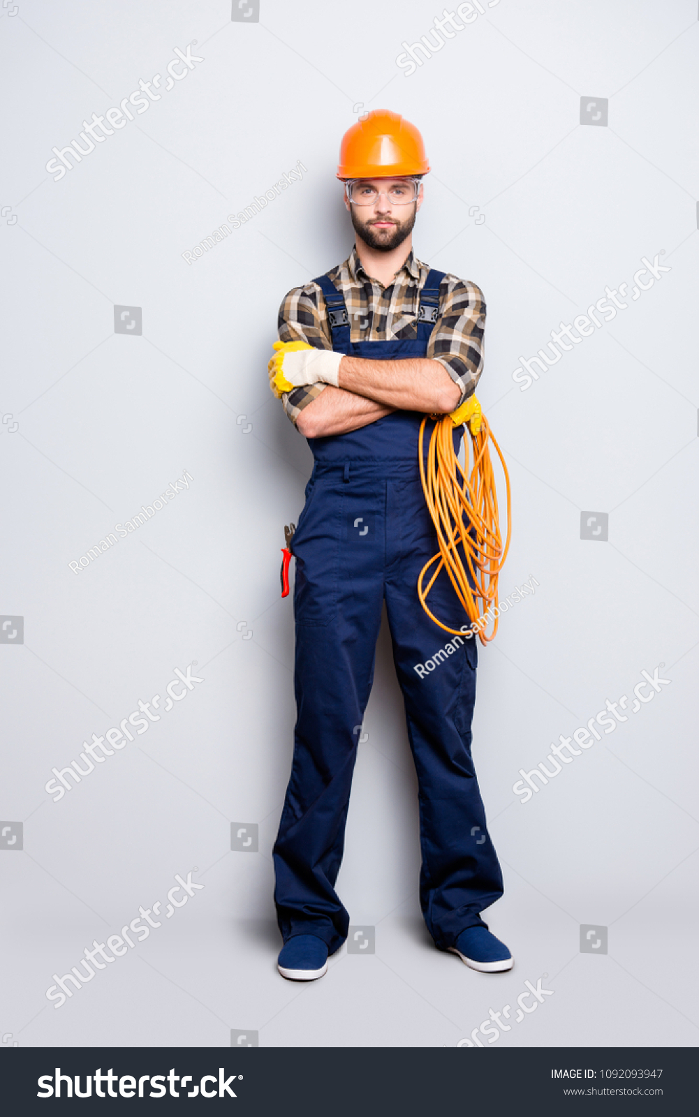 Full size body portrait of virile brutal electrician with bristle in overall, shirt, holding rolled cable in hand, having his arms crossed, looking at camera, isolated on grey background #1092093947