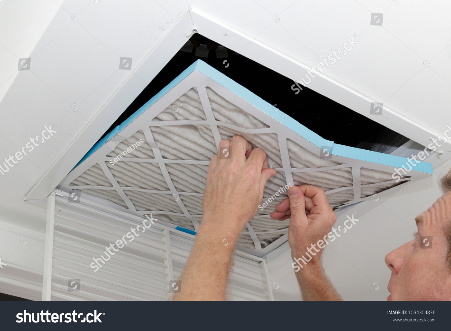 Person removing an old dirty air filter from a ceiling intake vent of a home HVAC system. Unclean gray square furnace air filter being taken out of a ceiling air vent. #1094304836