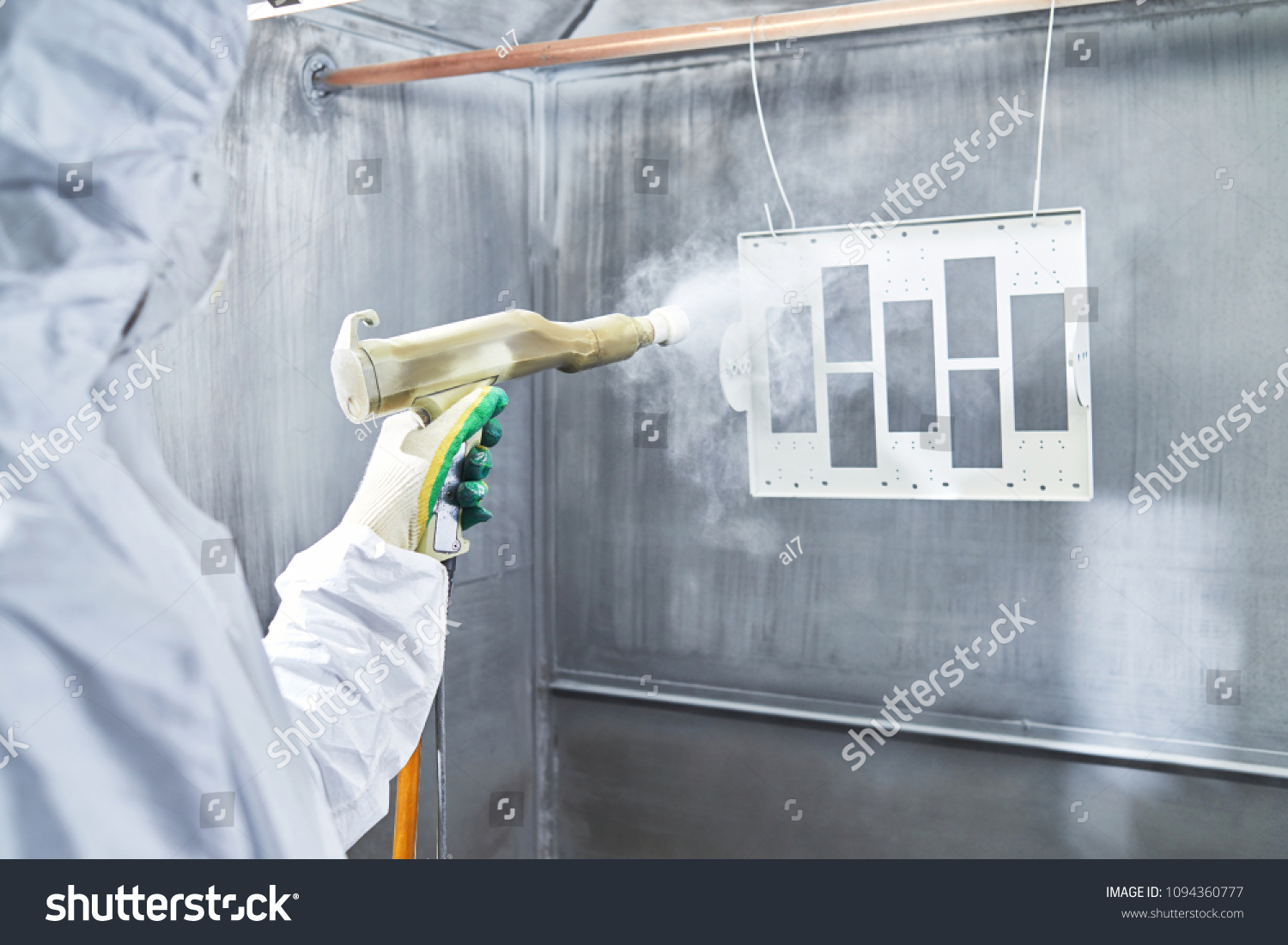 Powder coating of metal parts. Worker wearing protective wear performing powder coating of metal details in a special industrial camera. Hand holding powder coating sprayer #1094360777
