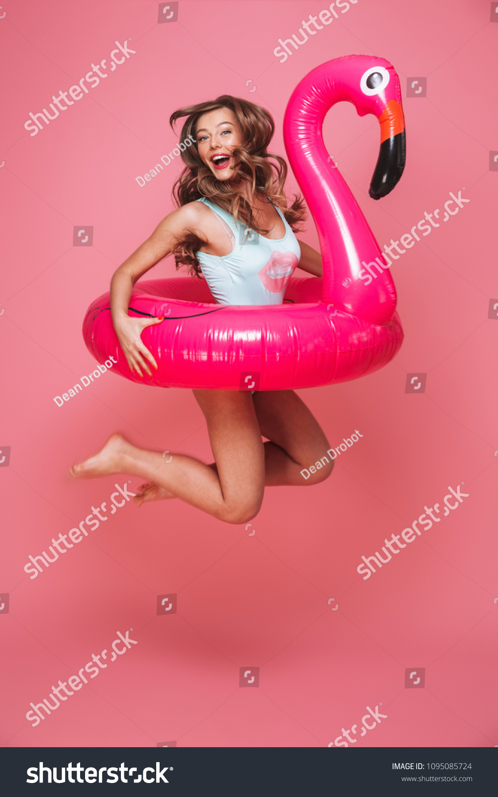 Full length portrait of a happy young woman dressed in swimsuit jumping with inflatable flamingo isolated over pink background #1095085724
