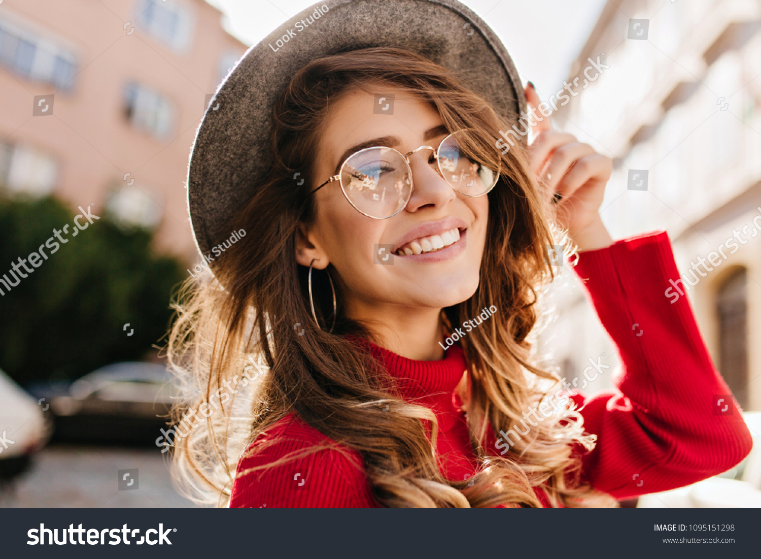 Close-up portrait of cheerful white woman in glasses touching her hat on blur background. Photo of fashionable girl with beautiful brown hair smiling to camera. #1095151298