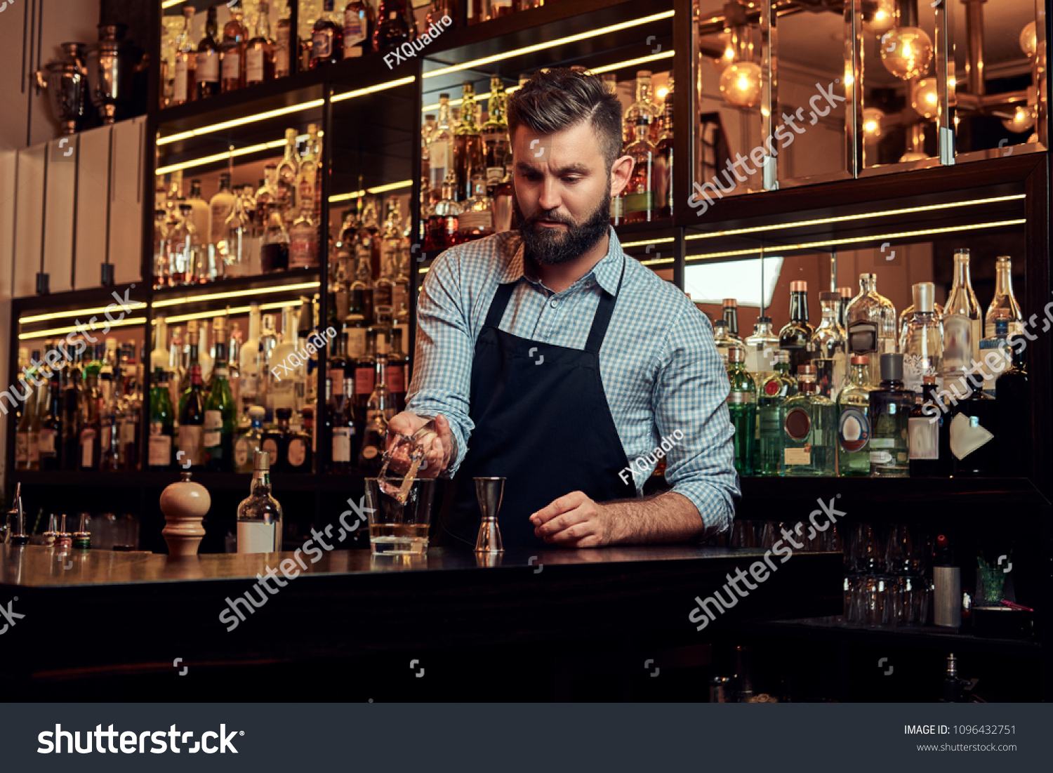 Stylish brutal barman in a shirt and apron makes a cocktail at bar counter background. #1096432751