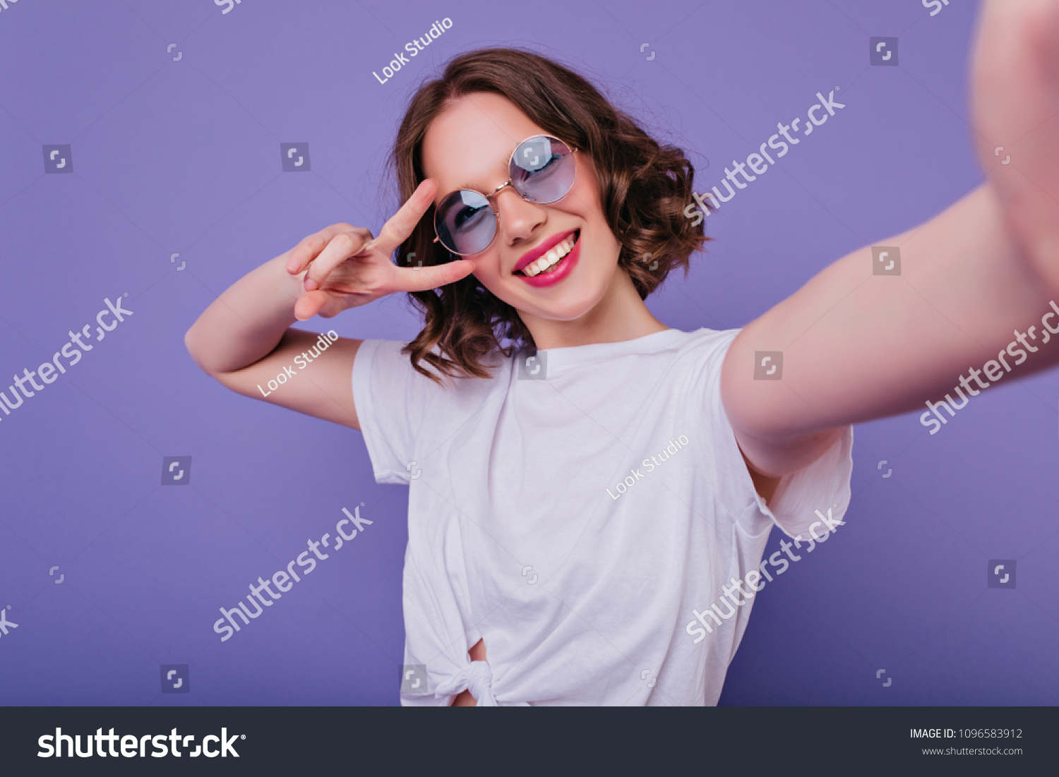 Pleasant girl with tattoo making selfie in studio and laughing. Good-looking young woman with brown wavy hair taking picture of herself on bright purple background. #1096583912
