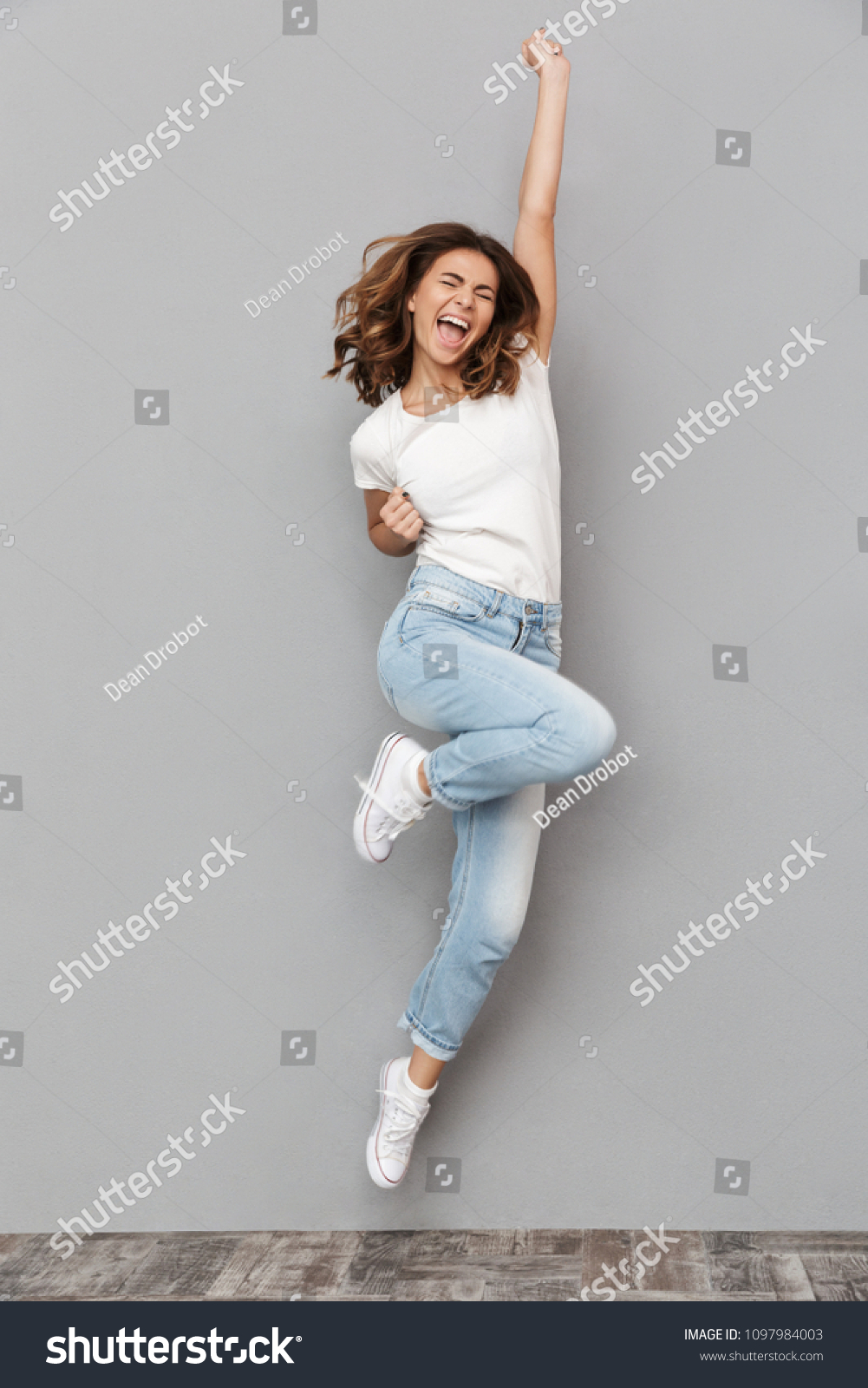 Full length portrait of a joyful young woman jumping and celebrating over gray background #1097984003