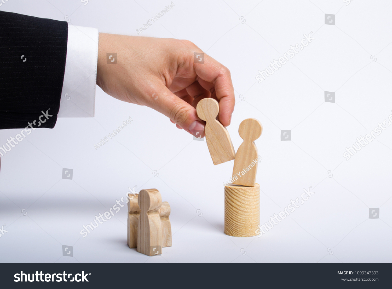The man's hand of a businessman in a business suit holds a wooden figure of a man in his hand and moves another figure from his post. The concept of firing an employee, the replacement of staff #1099343393