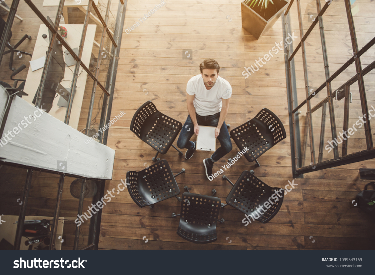 Top view full length portrait of serene male looking at camera while sitting near chairs and holding document in hands #1099543169
