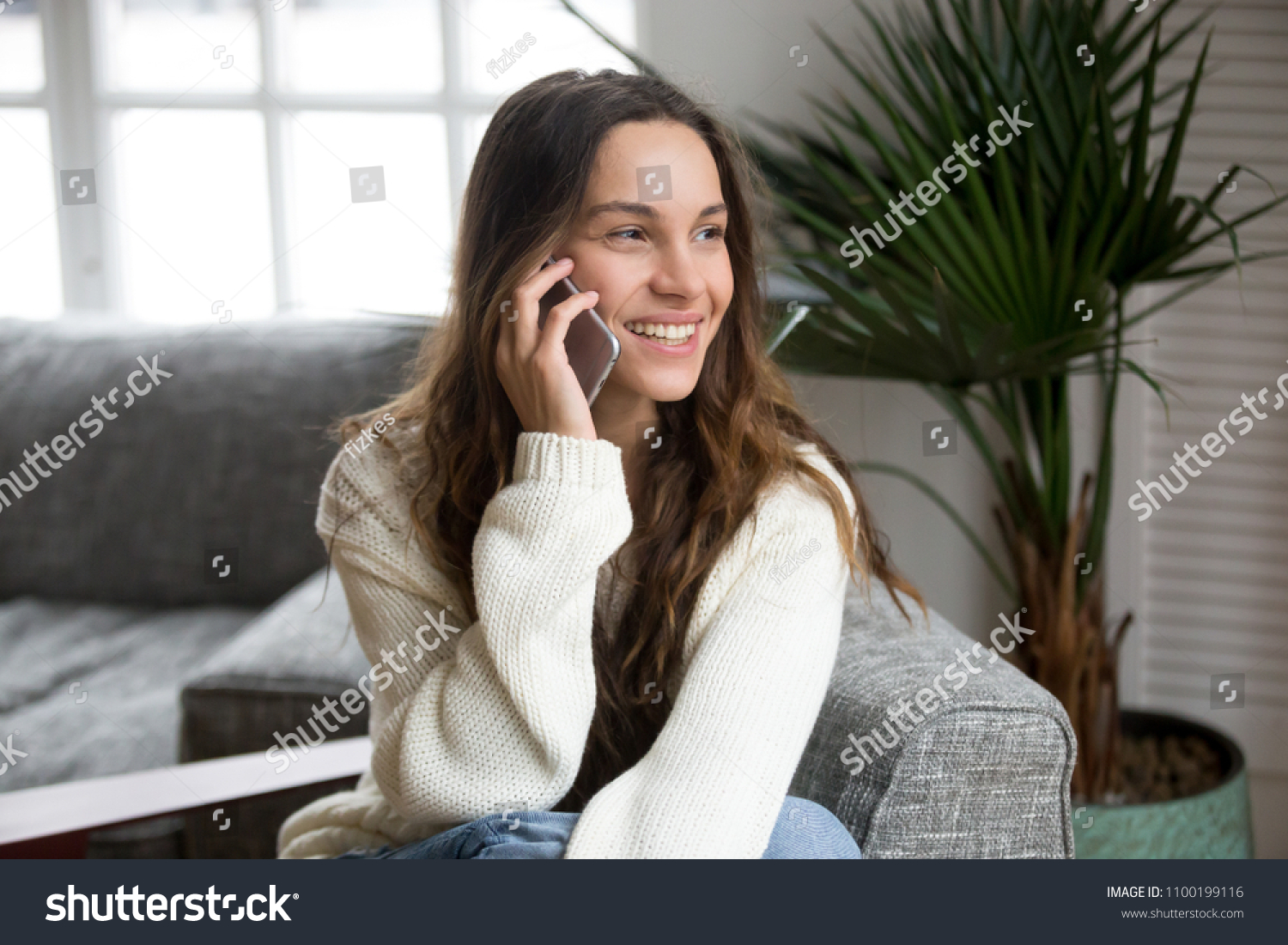 Smiling millennial woman talking on the phone at home, happy young girl holds cellphone making answering call, attractive teenager having pleasant conversation chatting by mobile with friend #1100199116