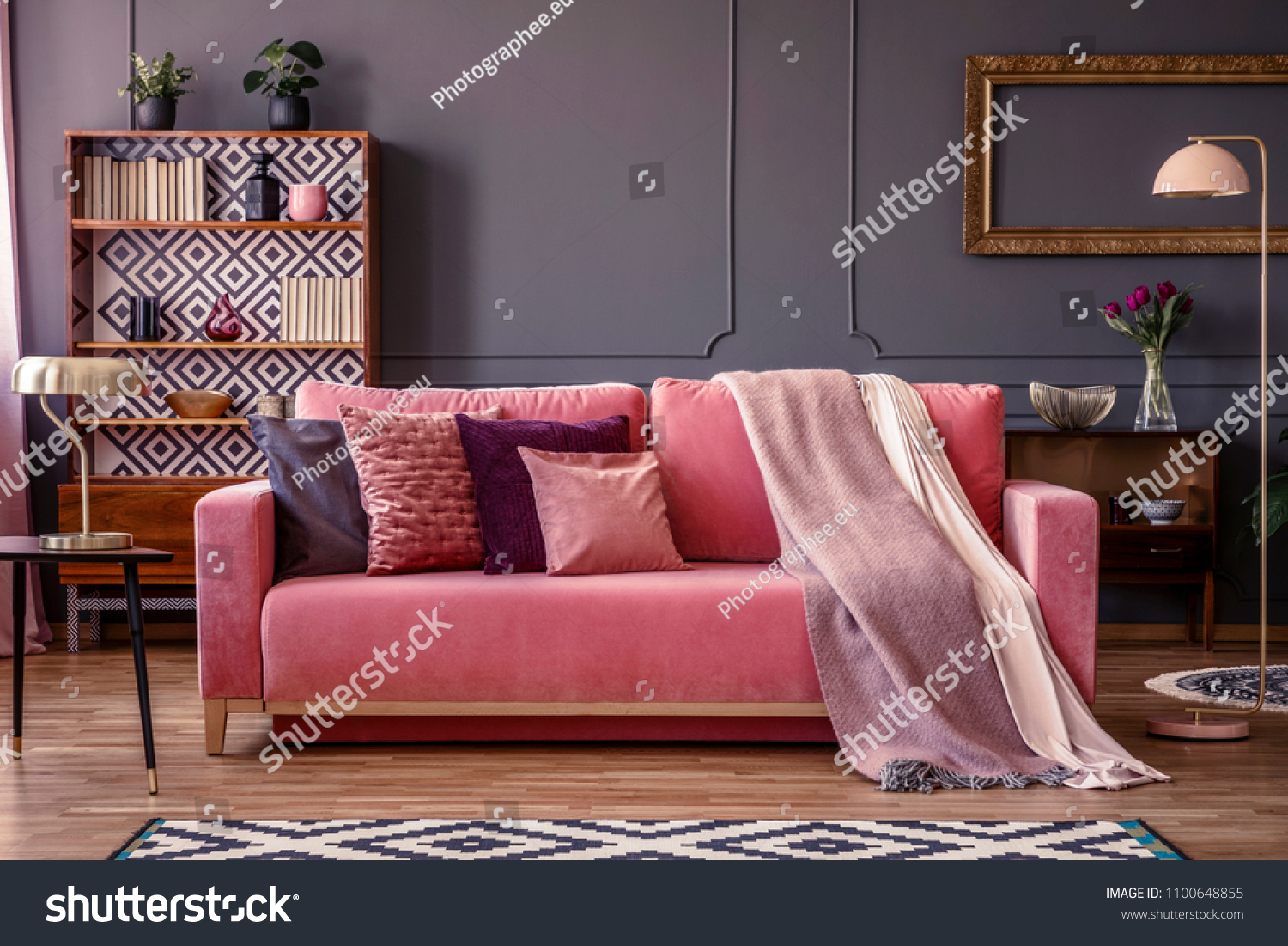 Front view of a pink sofa with pillows and blanket, vintage cupboard in the background in a glamorous living room interior #1100648855