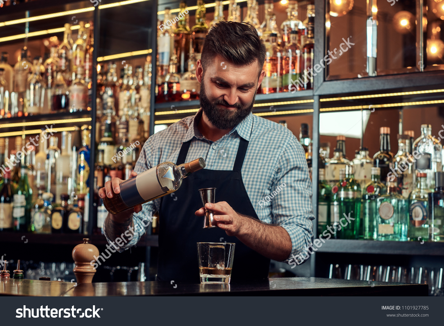 Stylish brutal barman in a shirt and apron makes a cocktail at bar counter background. #1101927785