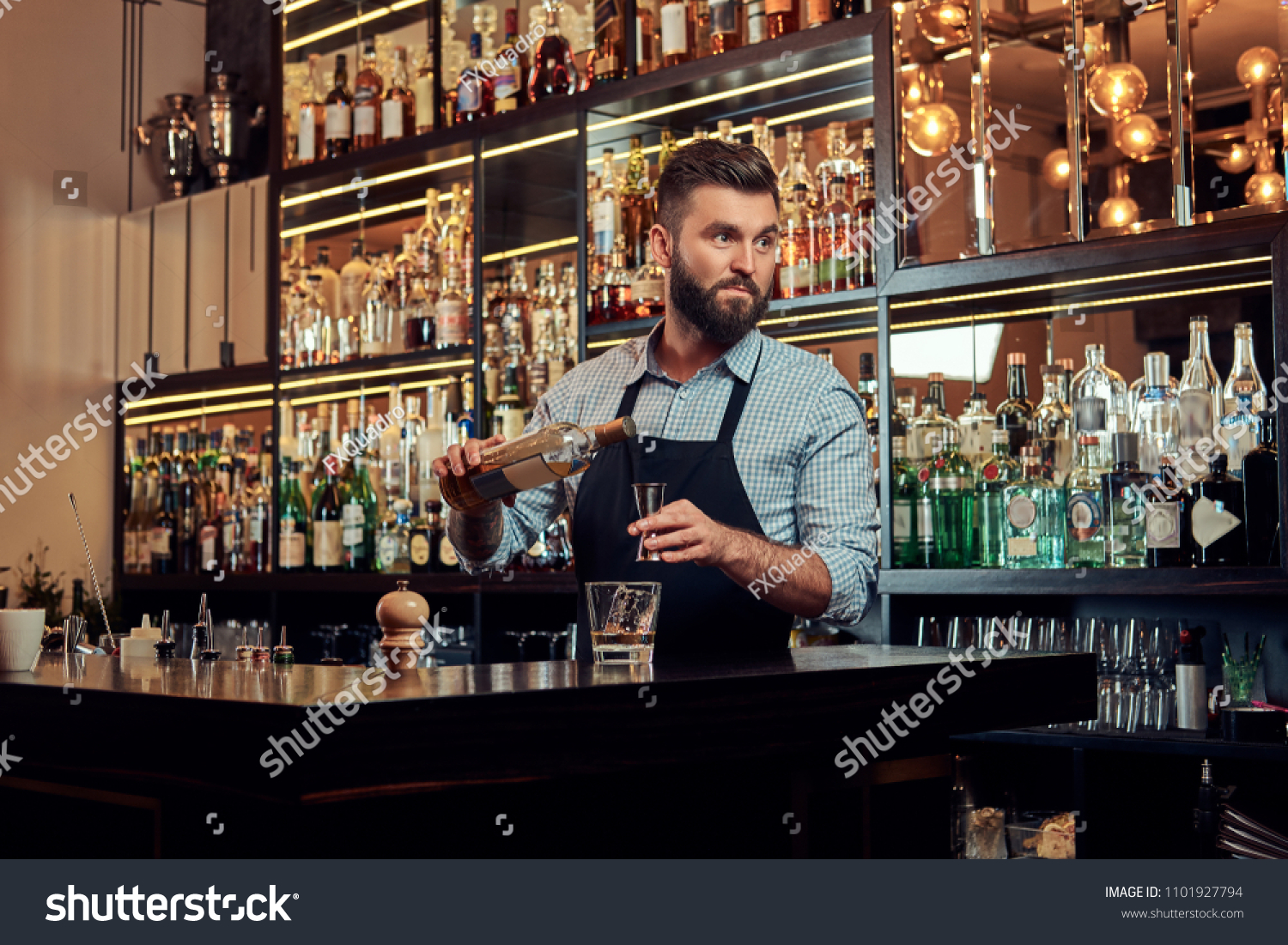 Stylish brutal barman in a shirt and apron makes a cocktail at bar counter background. #1101927794