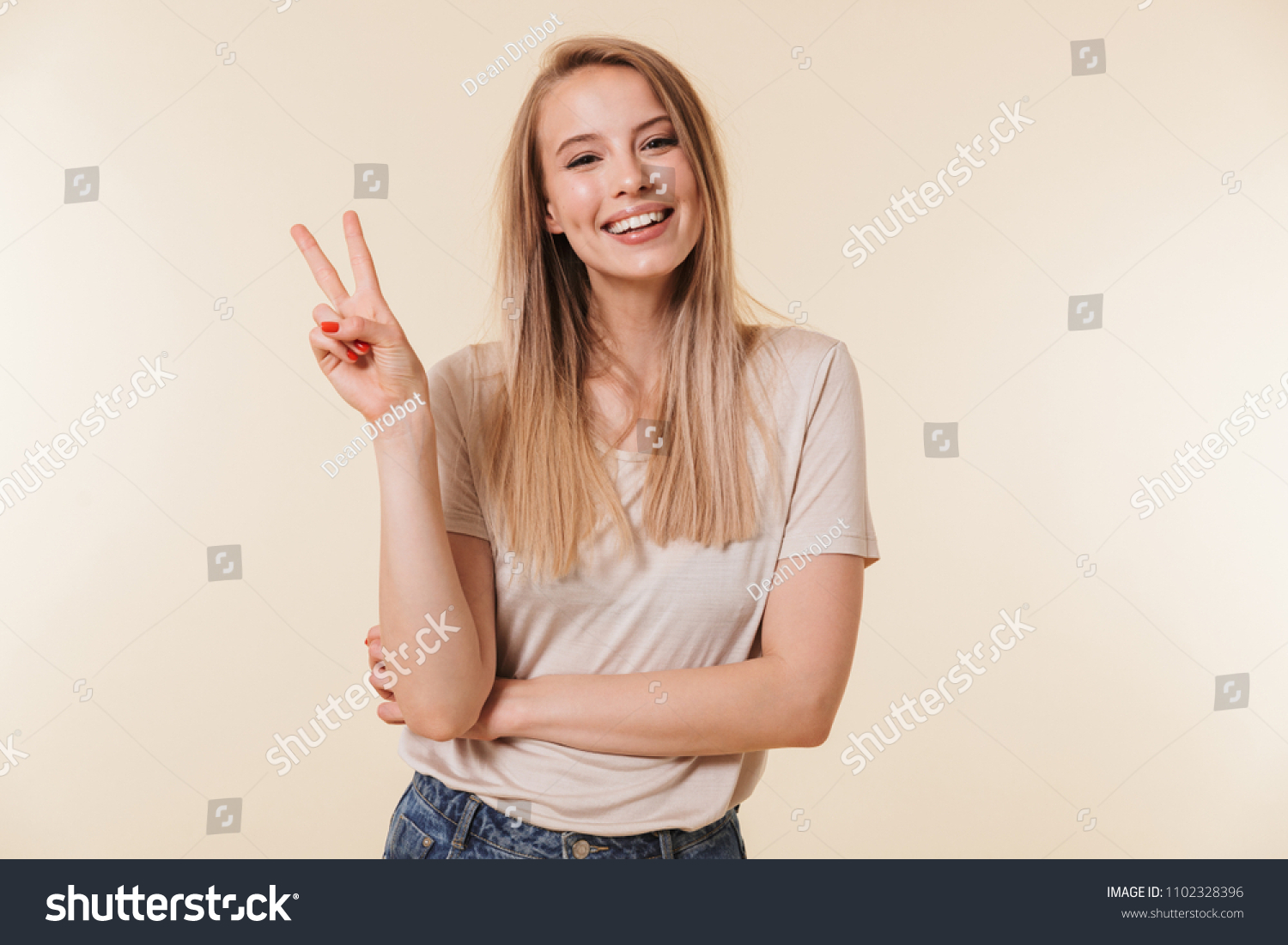Image of cheerful caucasian woman wearing casual clothing smiling and showing peace sign with two fingers isolated over beige background in studio #1102328396