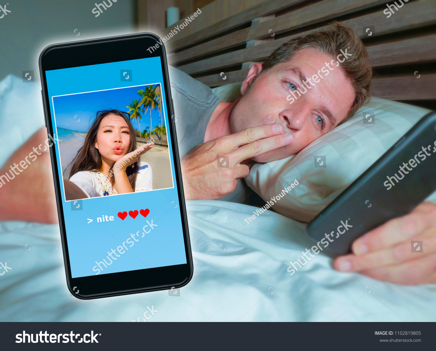 happy attractive man lying on bed sending good night kiss text with mobile phone having internet video call with Asian Chinese girlfriend in long distance relationship diversity couple concept  #1102819805