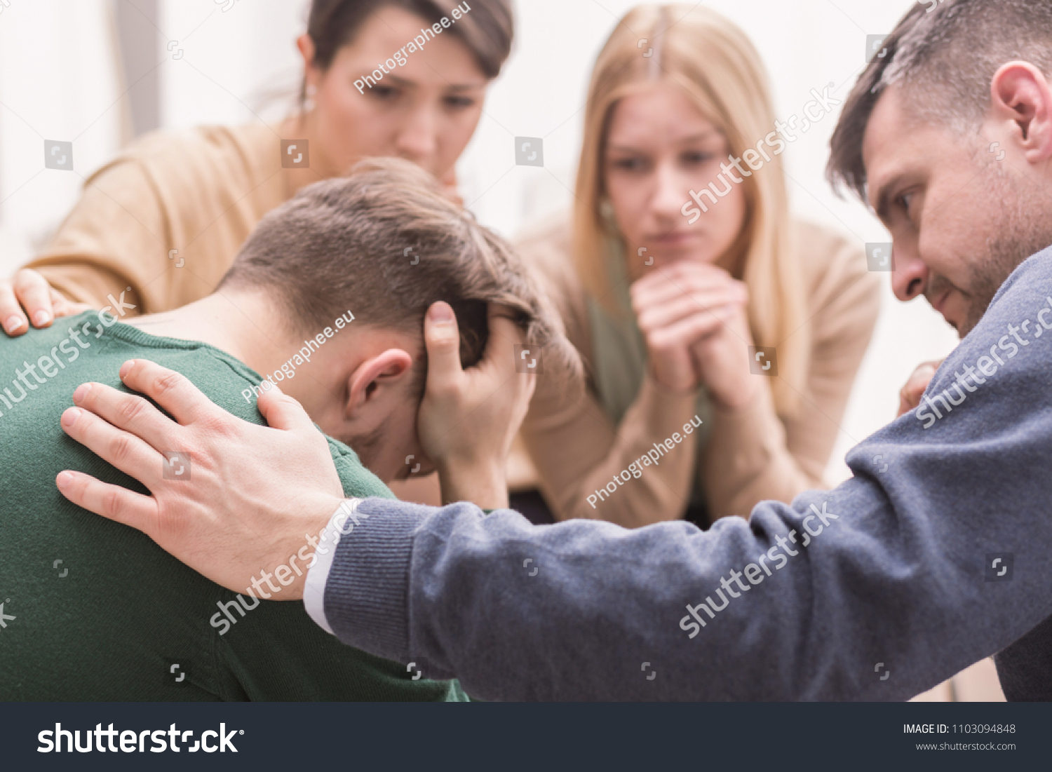 Close-up of a devastated young man holding his head in his hands and friends supporting him during group therapy #1103094848