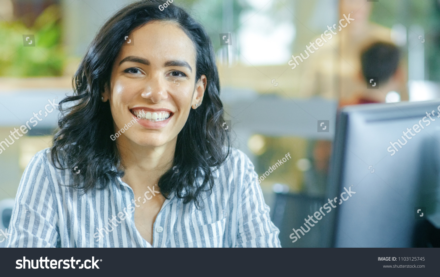 Portrait Shot of a Beautiful Young Hispanic Woman Working on a Computer, Smiling Warmly on the Camera. In the Background Busy Office with Working Colleagues. #1103125745