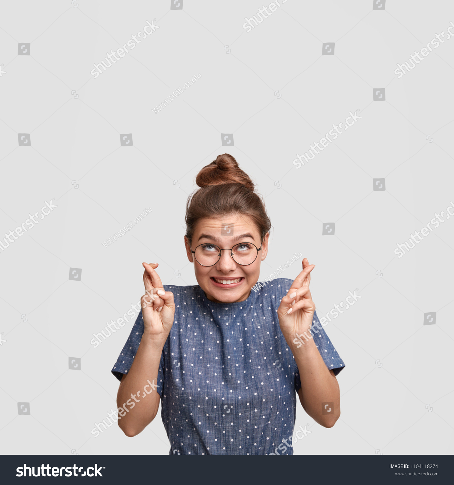 Vertical shot of wishful female sees falling star, crosses fingers as makes desirable wish, looks upwards, prays indoor against white background with blank space for your advertisement or text #1104118274