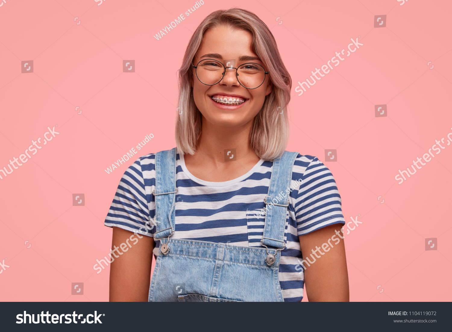 Glad smiling pretty female freelancer, has broad smile, demonstrates teeth with braces, wears casual t shirt with overalls, rejoices recieving reward, stands against pink background. Emotions concept #1104119072