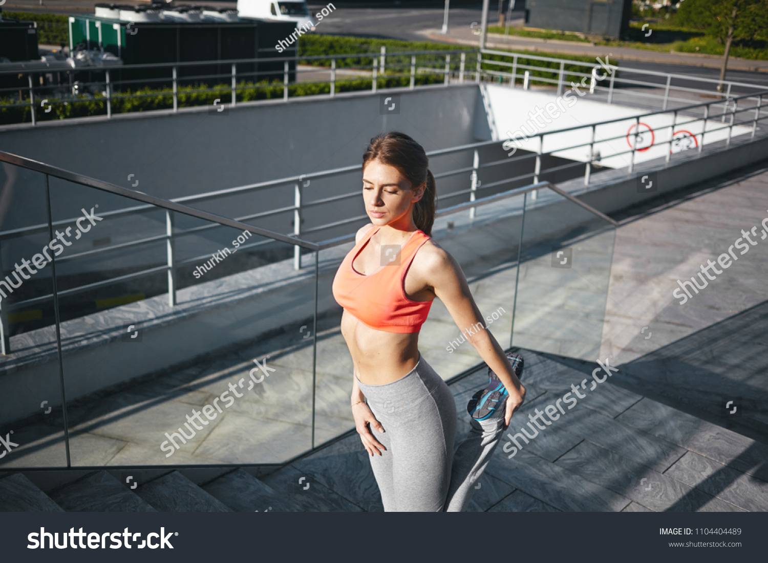Self determined young woman in trendy sportswear exercising outdoors in the morning before work, warming up muscles, preparing legs for running workout. Sports, cardio, strength and determination #1104404489